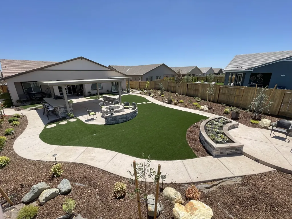 Aerial view of a backyard with a curved lawn, concrete walk, round fire pit court, raised planters, and a covered patio