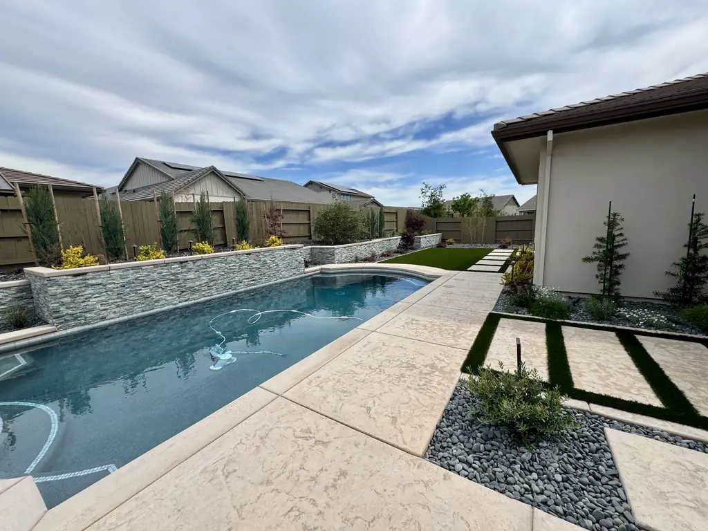 Long pool with a stone water wall, paved terrace, and planting beside a house