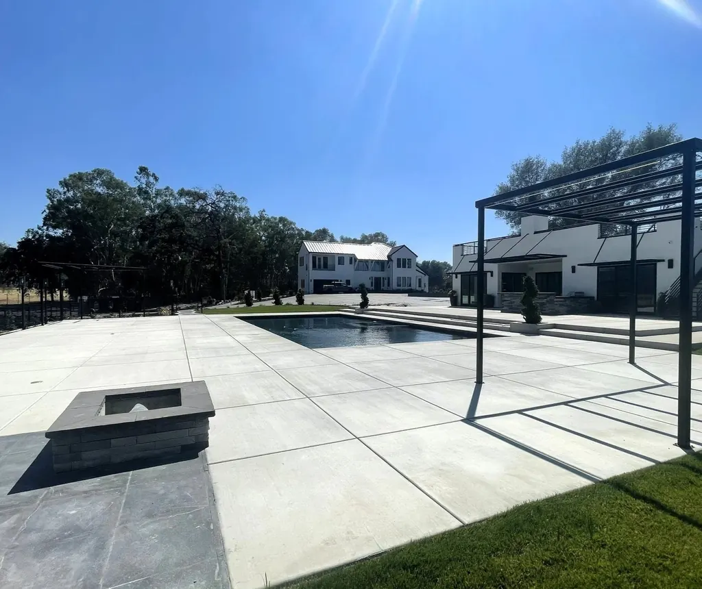 Wide concrete pool deck with a black steel pergola and square fire feature beside a long pool