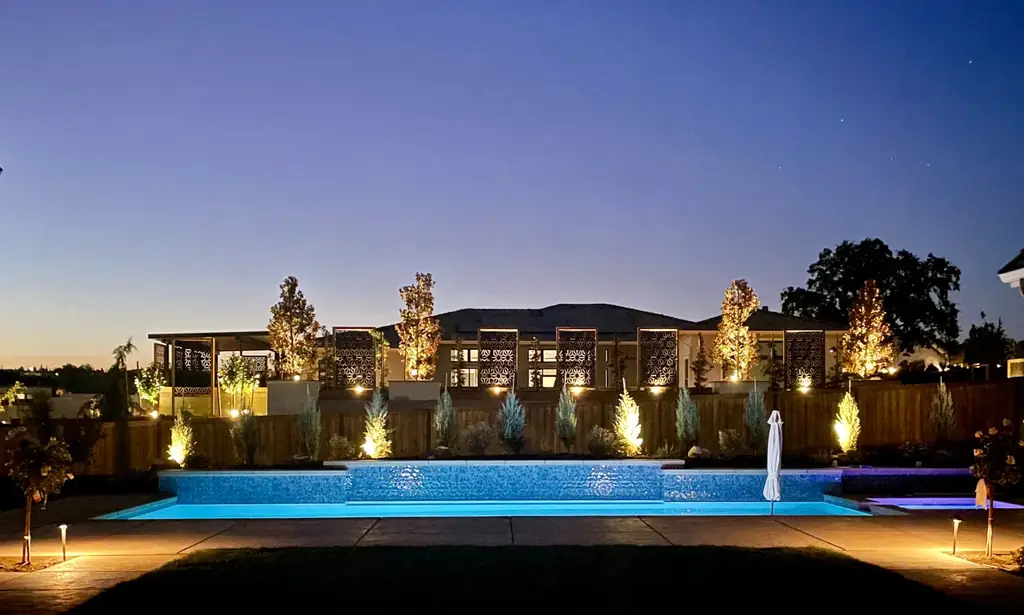 Pool and garden lit at dusk, with uplit trees and cypress along the fence line