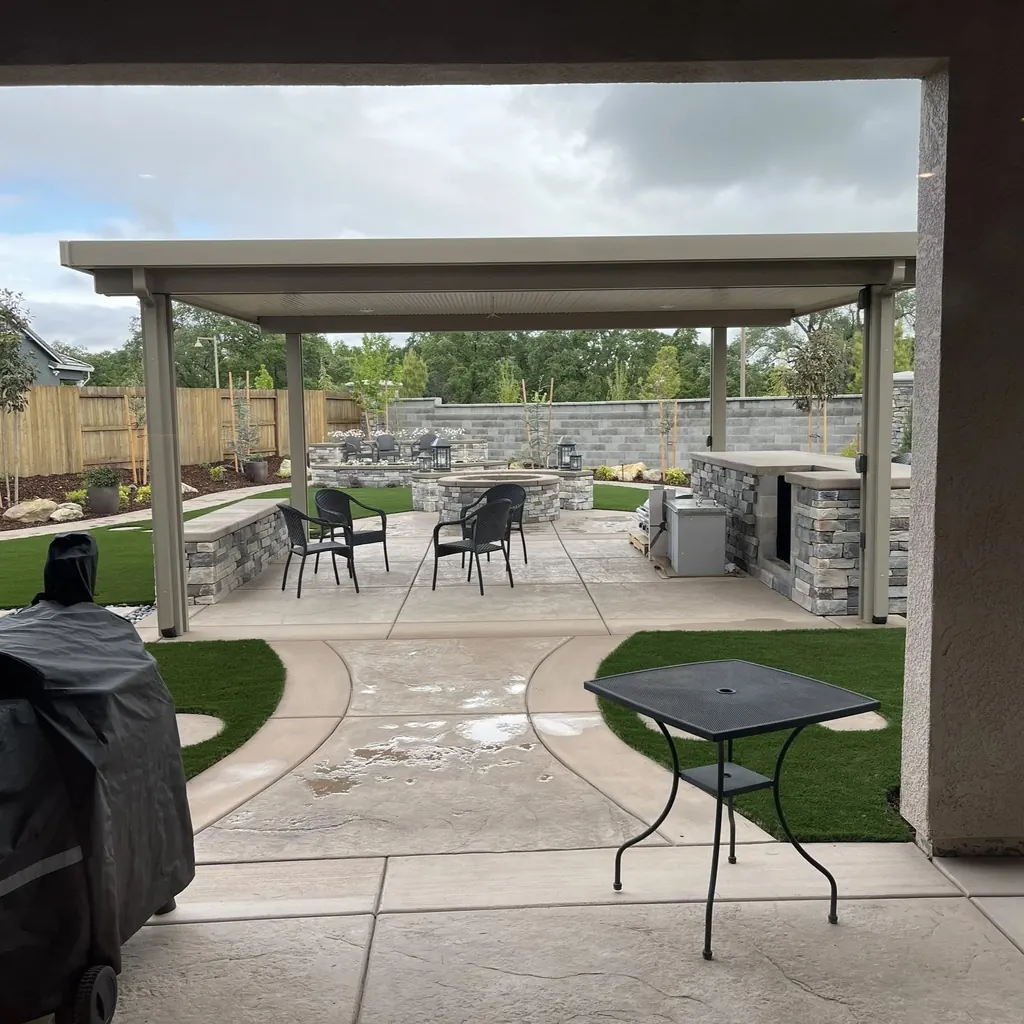 View from a covered porch to a patio cover with a stone outdoor kitchen, dining chairs, and turf