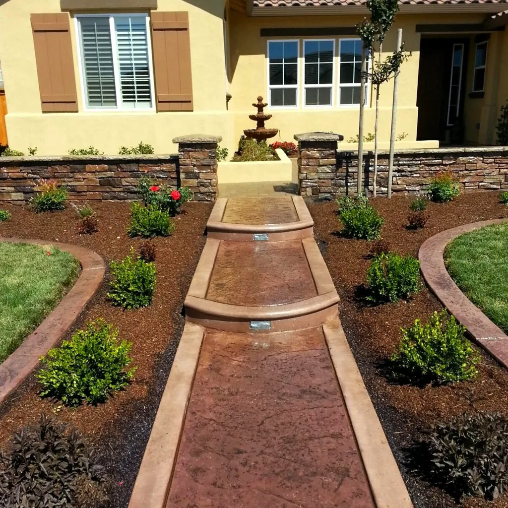 Two-tone stamped concrete walk leading to a tiered fountain between planting beds